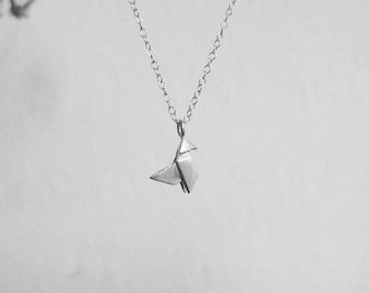 Może przedstawiać: Srebrzysty naszyjnik z małą, geometryczną zawieszką w kształcie ptaka przypominającą żurawia origami. Zawieszka wisi na delikatnym, cienkim metalowym łańcuszku z małymi, jednolitymi okrągłymi ogniwami, co nadaje całości prosty wygląd.