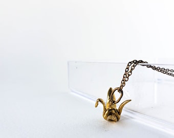 May include: A gold-toned metal pendant shaped like an origami crane suspended from a matching thin metal chain. The pendant features a textured, metallic finish and is attached to the chain by a small circular jump ring.