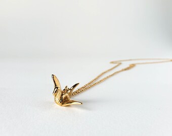 Puede incluir: Un collar de tono dorado que presenta un pequeño colgante de grulla de origami en tres dimensiones. El colgante está suspendido de una delicada cadena a juego de tono dorado que se extiende, mostrando un acabado metálico pulido.