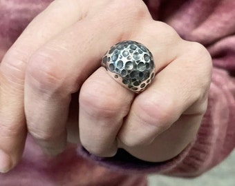 May include: A silver-toned ring featuring a prominent, rounded dome top with a textured, cratered surface. The metal has a darkened, oxidized finish within the recessed areas of the circular indentations, creating a high-contrast appearance.