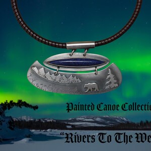 May include: A silver pendant necklace featuring a dark blue stone inlay and an engraved landscape scene with a bear and pine trees. The pendant is suspended from a dark brown cord. The text Painted Canoe Collection and Rivers To The West is visible.