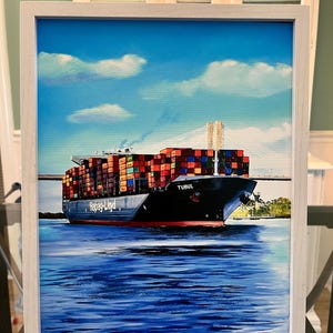 May include: A framed painting of a large cargo ship, the "TUBUL", sailing on blue water under a blue sky with fluffy white clouds. The ship is loaded with colorful shipping containers and is passing under a bridge. The painting is in a white frame.
