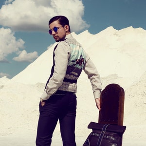 May include: A man in a light beige long-sleeve shirt and dark pants stands next to a dark brown wooden chair. He wears a dark vest with a light blue and white patterned back. A black bag with white text that reads 'KALK O KUBKU' is on the chair. The man wears sunglasses.