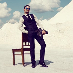 May include: A dark-blue suit vest and pants ensemble. The vest has a notched lapel and is worn over a white long-sleeved shirt and dark-colored tie. A brown leather briefcase is held in one hand. The man is wearing blue sunglasses.