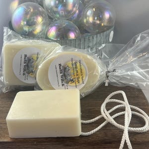 May include: Two bars of white soap with a yellow label that reads "Bath Witch" and "Soap on a Rope" with a white rope attached to one of the bars. The soap is packaged in clear plastic.