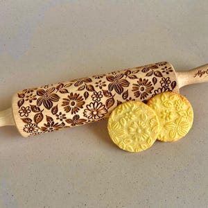 Può includere: Un mattarello di legno con un motivo floreale e due biscotti goffrati. Il mattarello è marrone chiaro con un motivo ripetuto di fiori e foglie. I biscotti sono rotondi e presentano un motivo floreale simile.