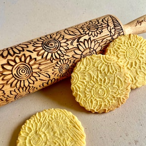 May include: A wooden rolling pin with a sunflower design engraved into the surface. The rolling pin is used to create decorative patterns on cookies. Three cookies with sunflower designs are shown in the image.
