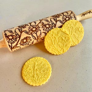 Puede incluir: Un rodillo de madera con un diseño floral de girasoles y hojas. El rodillo se utiliza para crear diseños en relieve en las galletas. Se muestran tres galletas con el mismo diseño de girasol junto al rodillo.
