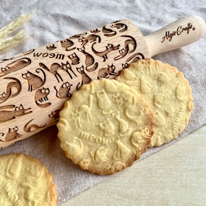 Puede incluir: Un rodillo de madera con un diseño de gatos y el texto "wosm" junto a varias galletas redondas con diseños de gatos y la palabra "meow". El rodillo tiene el texto "Algis Crafts".