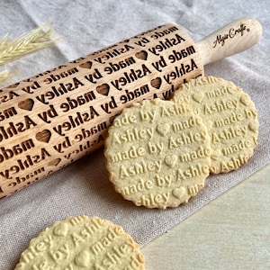 May include: A wooden rolling pin with the name "Ashley" and heart shapes engraved on it, alongside three cookies stamped with "made by Ashley". The rolling pin has a light wood finish, and the cookies are round.