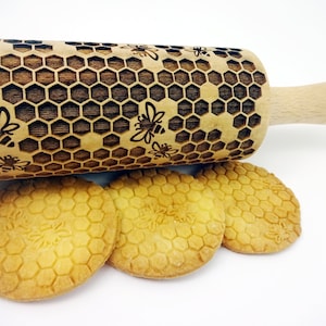 May include: A wooden rolling pin with a honeycomb and bee design. The rolling pin is used to create embossed patterns on cookies. Three cookies with the honeycomb pattern are shown in the image.