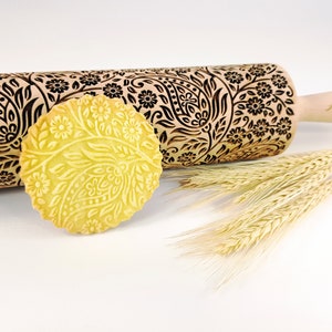 May include: A light wood rolling pin with a floral and paisley pattern, and a round biscuit with the same embossed design. The rolling pin has a handle with the text "Alina Crafts". Wheat stalks are placed next to the biscuit.