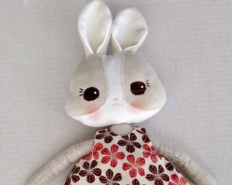 May include: A handmade fabric bunny doll wearing a white dress with a brown and orange floral pattern. The doll has a white face with brown eyes and a pink blush.