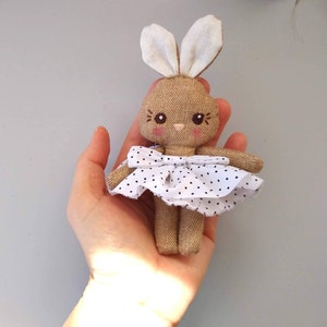 Puede incluir: Una pequeña muñeca de conejo de tela marrón que lleva un vestido blanco con lunares negros y un lazo blanco. La muñeca tiene ojos bordados y un rubor rosado.