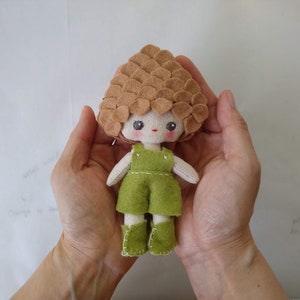 May include: A small felt doll with a brown felt hair, green dress, and brown felt boots. The doll is being held in the palms of two hands.