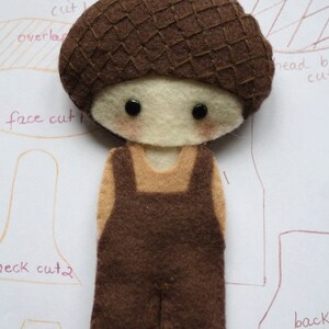 May include: A felt doll with a brown acorn hat, brown overalls, and a tan shirt. The doll has a white face with brown eyes and rosy cheeks. The doll is on a white background with handwritten instructions for making the doll.