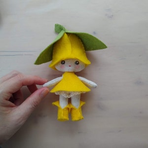 May include: A handmade felt doll in the shape of a yellow flower with a green leaf hat. The doll has a yellow dress with a lace trim and yellow boots. The doll's face is hand-stitched with simple features.