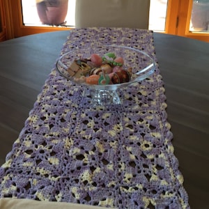 May include: A purple and white crocheted table runner with a glass bowl filled with colorful Easter eggs on a dark wood table.