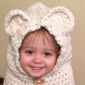 May include: A white crocheted hooded hat with bear ears for a toddler. The hat has a button closure and is made with a soft, fluffy yarn.