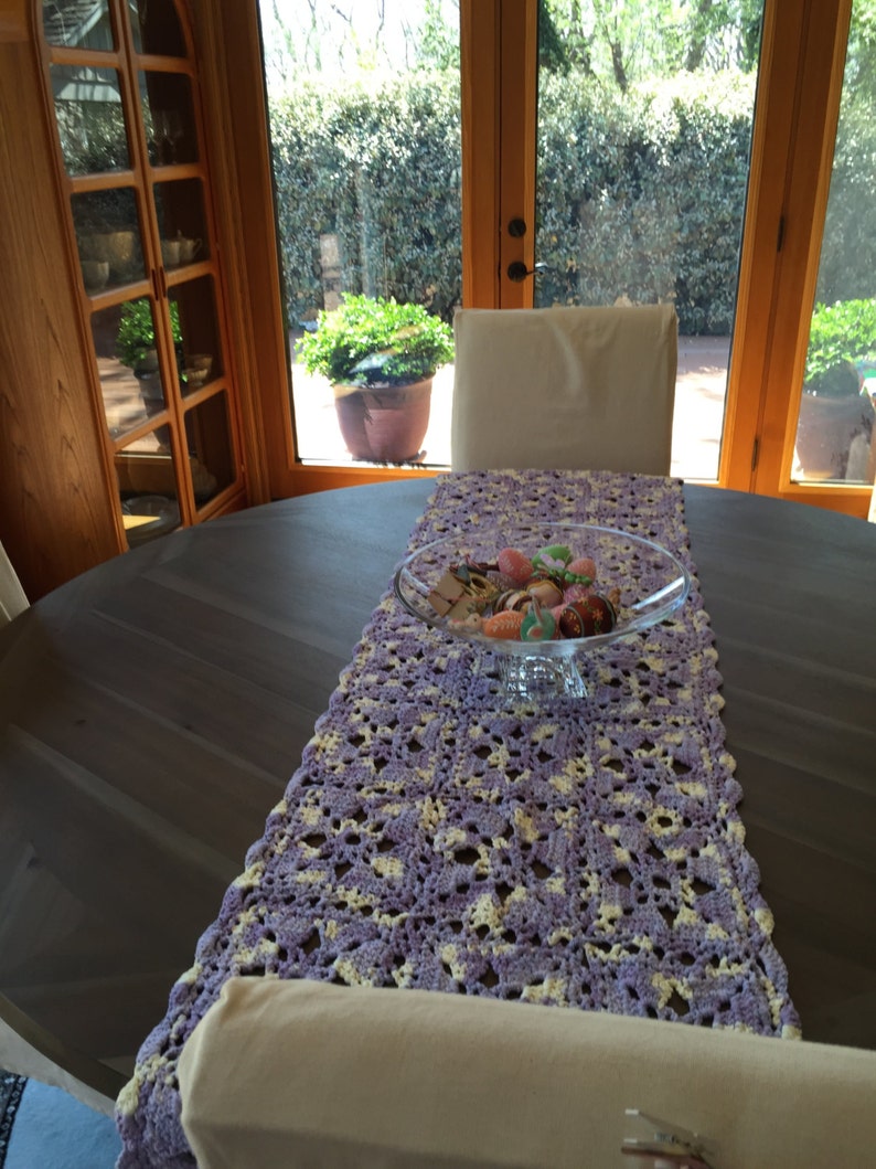 May include: A purple and white crocheted table runner with a glass bowl filled with colorful Easter eggs on a dark wood table.