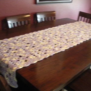 May include: A crocheted table runner in shades of purple and yellow, laid on a dark wood table with chairs.