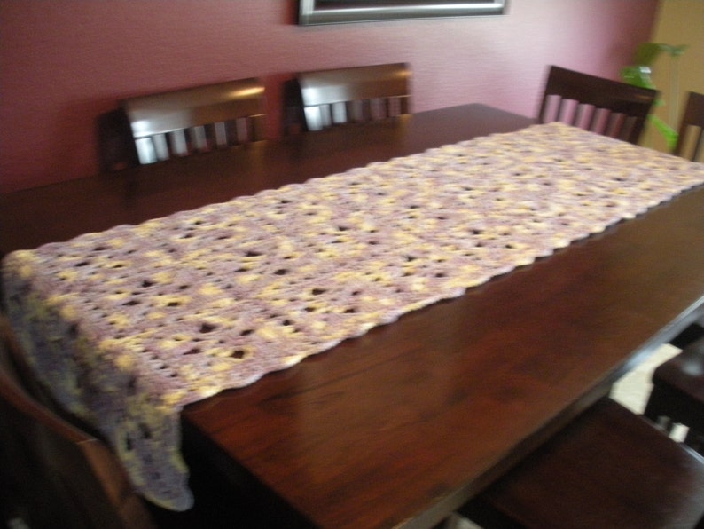 May include: A crocheted table runner in shades of purple and yellow, laid on a dark wood table with chairs.