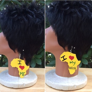 Puede incluir: Un par de pendientes de madera amarilla con forma del continente africano. Los pendientes tienen las palabras "Me encanta mi pelo" escritas en verde y rojo.