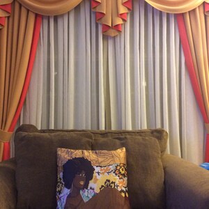 Puede incluir: Un sof&aacute; marr&oacute;n con un coj&iacute;n que presenta una ilustraci&oacute;n colorida de una mujer con piel oscura y cabello rizado. El sof&aacute; est&aacute; frente a una ventana con cortinas transparentes y una cenefa decorativa.