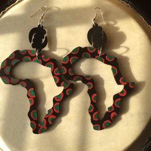 Puede incluir: Un par de pendientes colgantes con forma del continente africano. Los pendientes están hechos de madera y pintados de negro con círculos rojos y verdes. Los pendientes tienen un gancho negro y plateado.