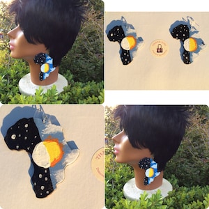 Puede incluir: Un par de pendientes con forma del continente africano, pintados con un cielo nocturno en un lado y un sol y una luna en el otro. Los pendientes están hechos de madera y están pintados en negro, blanco, azul, amarillo y naranja.