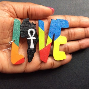 Puede incluir: Letras de madera que deletrean "LOVE" con un contorno de África en el medio. Las letras están pintadas en varios colores, incluyendo verde, naranja, rojo, amarillo y azul. El contorno de África es negro con un símbolo Ankh blanco.