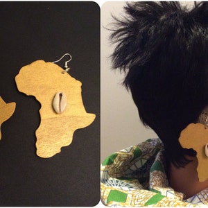 Puede incluir: Un par de pendientes de madera pintados de oro con forma del continente africano, cada uno con una pequeña concha de cauri blanca en el centro.