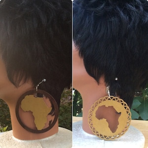 Puede incluir: Un par de pendientes de madera con un contorno dorado del continente africano. Los pendientes son redondos y tienen un fondo de madera marrón.