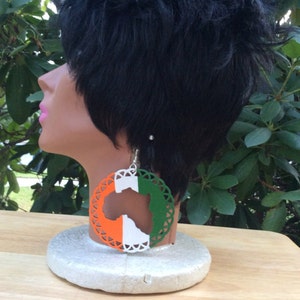 Puede incluir: Un par de pendientes grandes y redondos con un diseño blanco, naranja y verde. Los pendientes presentan un recorte del continente africano.