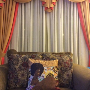Puede incluir: Un sof&aacute; marr&oacute;n con tres almohadas. La almohada del centro presenta un dise&ntilde;o floral colorido con la cara de una mujer negra en el centro. El sof&aacute; est&aacute; delante de una ventana con cortinas blancas transparentes y una cenefa dorada y roja.