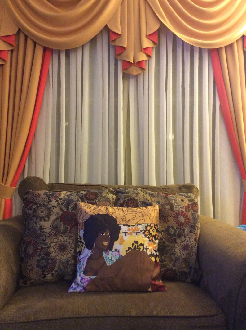 Puede incluir: Un sof&aacute; marr&oacute;n con tres almohadas. La almohada del centro presenta un dise&ntilde;o floral colorido con la cara de una mujer negra en el centro. El sof&aacute; est&aacute; delante de una ventana con cortinas blancas transparentes y una cenefa dorada y roja.