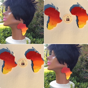 Puede incluir: Un par de pendientes de madera con forma del continente africano. Los pendientes están pintados en un degradado de naranja, amarillo y rojo. Los pendientes se muestran en una cabeza de maniquí.