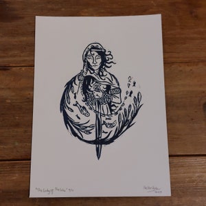 May include: A blue and white linocut print of a woman holding a sword, surrounded by fish and reeds. The print is titled "The Lady of the Lake" and is numbered 5/10.