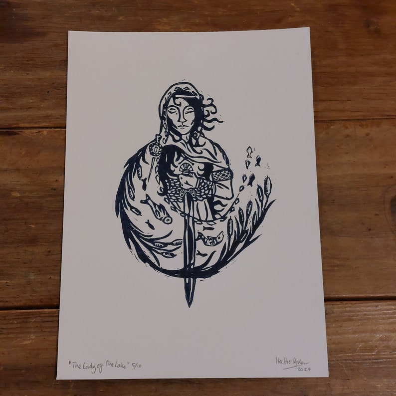 May include: A blue and white linocut print of a woman holding a sword, surrounded by fish and reeds. The print is titled "The Lady of the Lake" and is numbered 5/10.