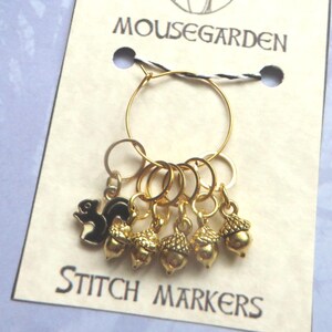 May include: A set of six gold stitch markers featuring a squirrel and acorns. The markers are attached to a gold ring and are labelled "MOUSEGARDEN STITCH MARKERS".