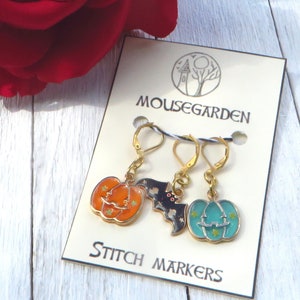 May include: Set of three Halloween-themed stitch markers. The markers feature a black bat, an orange pumpkin, and a teal pumpkin. Each marker has a gold loop and a gold clasp.