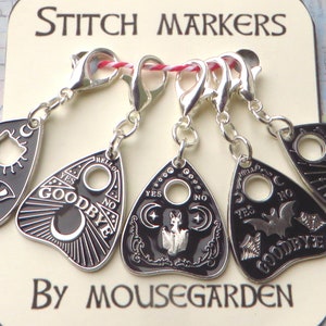 May include: Set of five black and silver stitch markers shaped like a Ouija board. Each marker features a different design, including a cat, a bat, a fox, and a crescent moon. The markers are labeled with the words "YES", "NO", "GOODBYE", and "HELLO". The text "STITCH MARKERS" and "BY MOUSEGARDEN" are printed on a white card behind the markers.