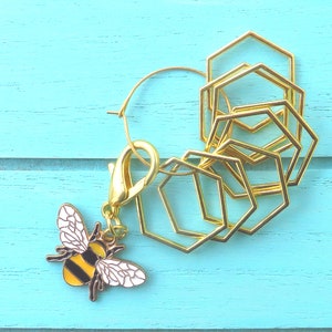 May include: A gold-coloured bee charm with black and yellow stripes and white wings, attached to a gold hoop and several gold geometric shapes. The items are on a light blue wooden surface.