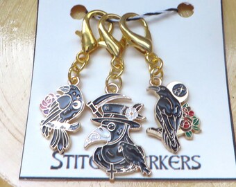 May include: Set of three gold-toned stitch markers with black and gold enamel designs. Each marker features a different gothic-themed charm: a raven, a plague doctor, and a raven with a rose. The word "STITCH MARKERS" is printed on the card.
