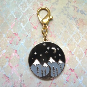 May include: Gold-toned metal keychain with a round charm featuring a black and white illustration of mountains, a crescent moon, and stars against a dark blue sky.