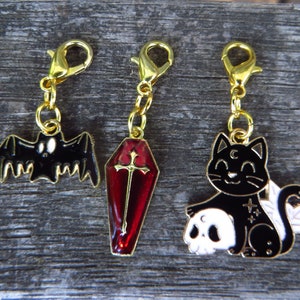 May include: Three gold-tone charms with Halloween themes. The first charm is a black bat with white eyes. The second charm is a red coffin with a gold cross. The third charm is a black cat holding a white skull with a crescent moon on its forehead.