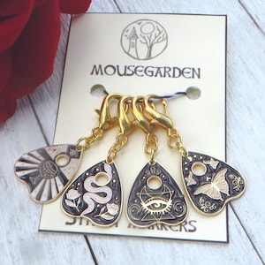 Peut inclure: Ensemble de quatre marqueurs de points de suture dorés en forme de cœurs de planchette Ouija noirs et blancs. Chaque marqueur présente un motif différent, notamment un serpent, un œil, un papillon et un soleil avec un verre de vin. La carte indique "Mousegarden Stitch Markers".