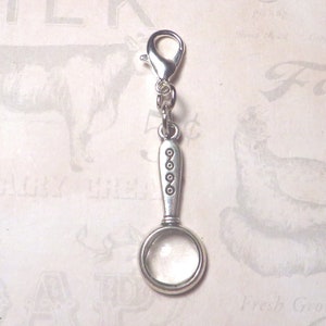 May include: Silver charm featuring a magnifying glass design. The charm has a silver loop for attaching to a bracelet or necklace. The magnifying glass has a clear glass lens and a silver handle with decorative details.