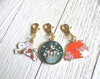 May include: Three enamel charms with gold-coloured clasps. One charm is a white rabbit with red roses, another is a teal circle with mushrooms and a butterfly, and the last is an orange fox with pink flowers.