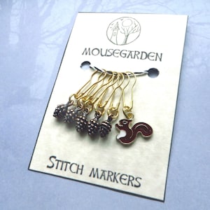 Peut inclure: Ensemble de 7 marqueurs de point en or avec des breloques en forme de gland et d'écureuil. Les breloques sont dans des tons de brun et d'or. La carte indique "Mousegarden Stitch Markers".